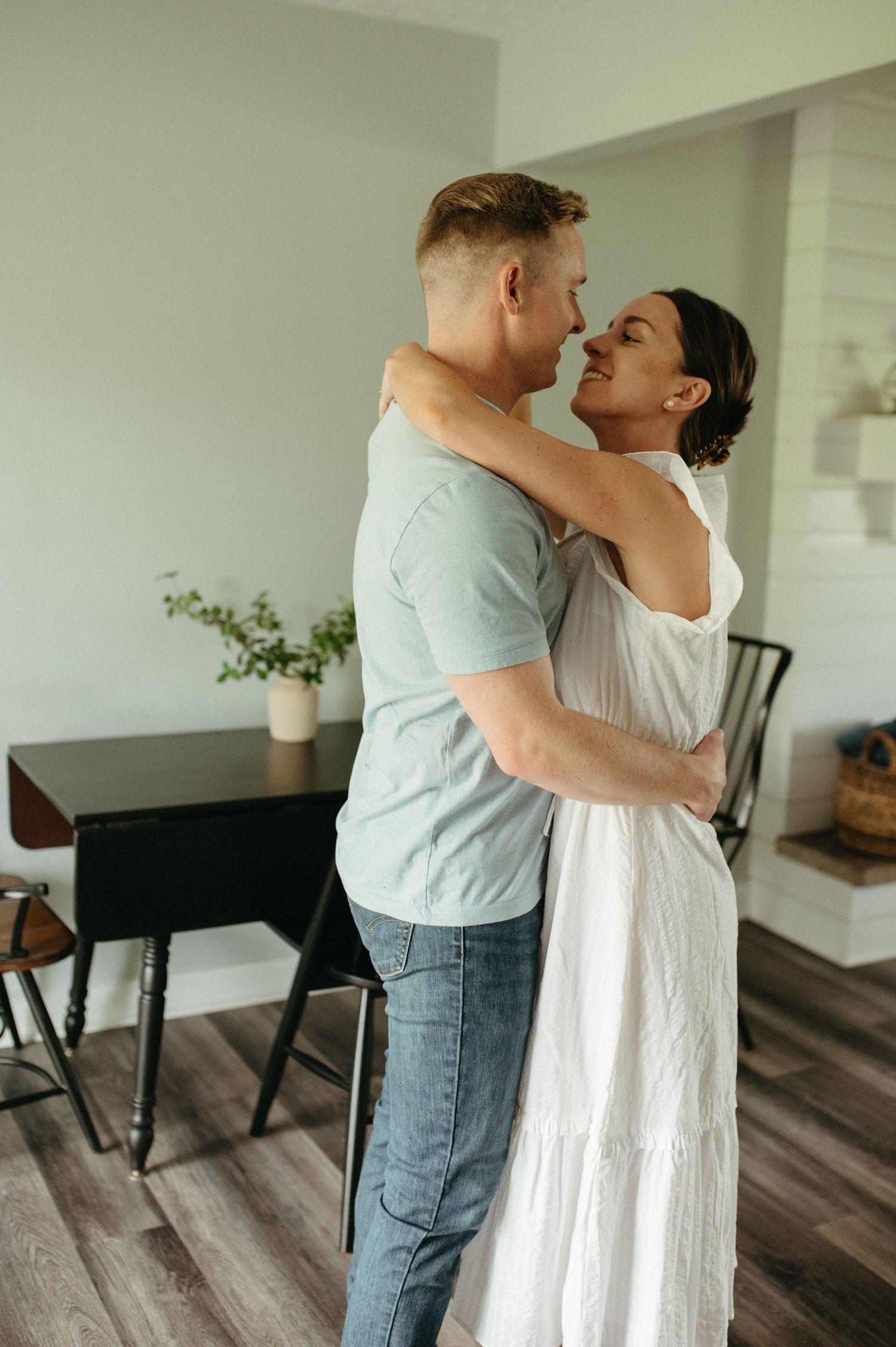 A couple embraces while standing in their home, she in a white dress and he in casual attire.