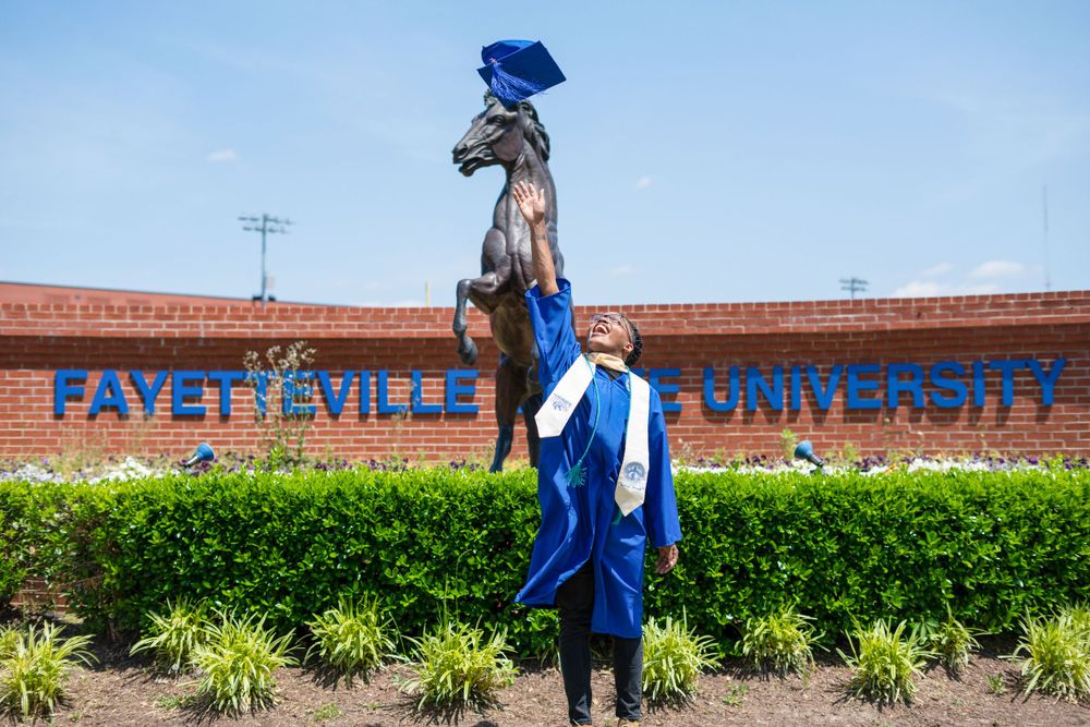 Person in blue graduation attire walks along brick wall with landscaping.