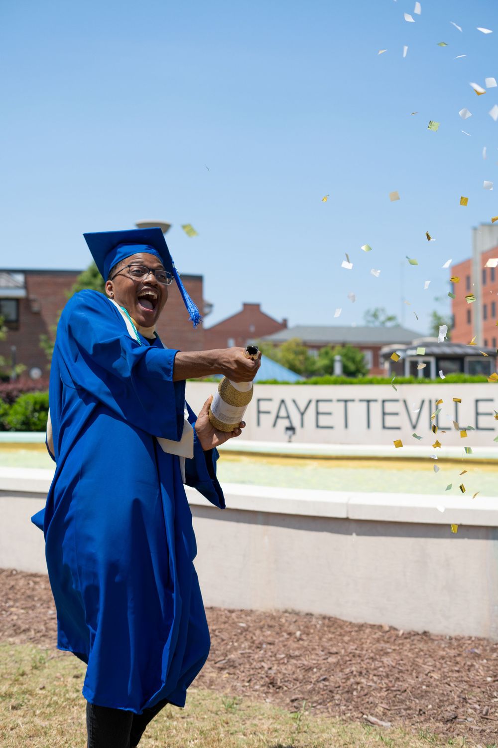 Graduate in blue cap and gown celebrates with confetti flying in front of Fayetteville sign.