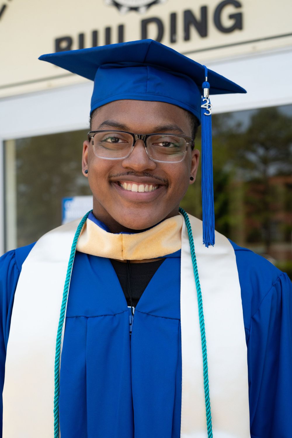 Graduate wearing blue cap and gown with white stole and green cord poses for photo.