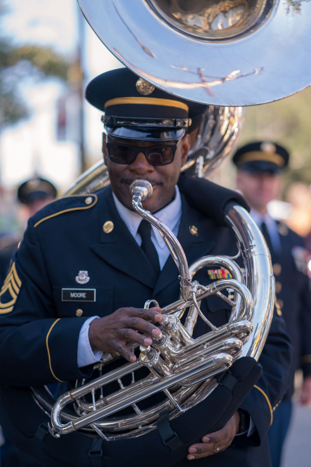Military band members play brass instruments during an outdoor performance.