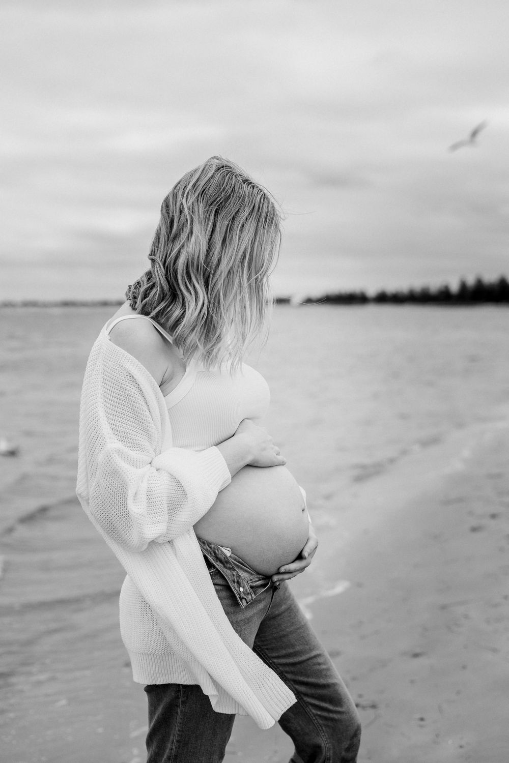 Black and white photo of pregnant woman standing on beach holding belly.