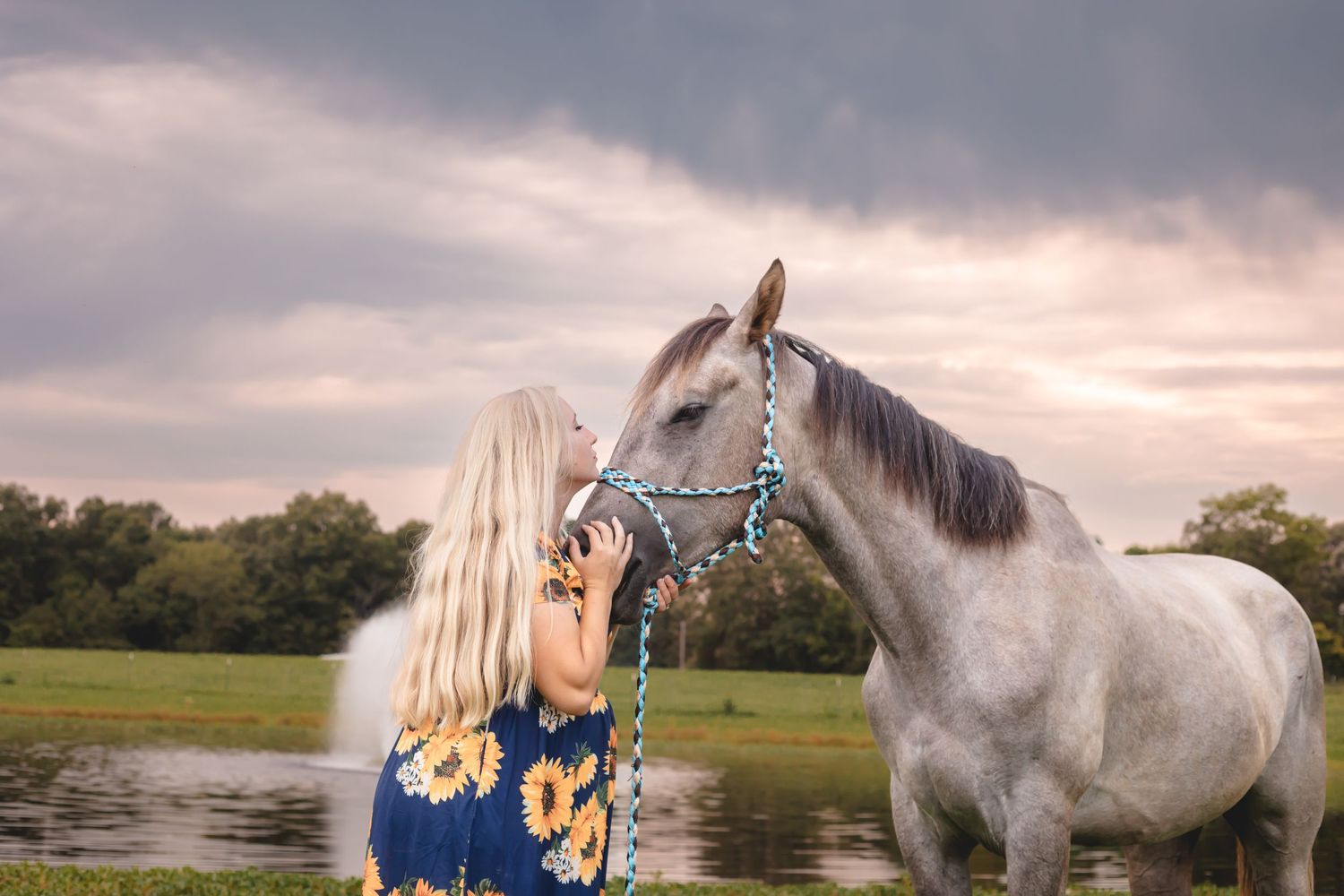 White horse and person in blue dress stand together by a pond at sunset.