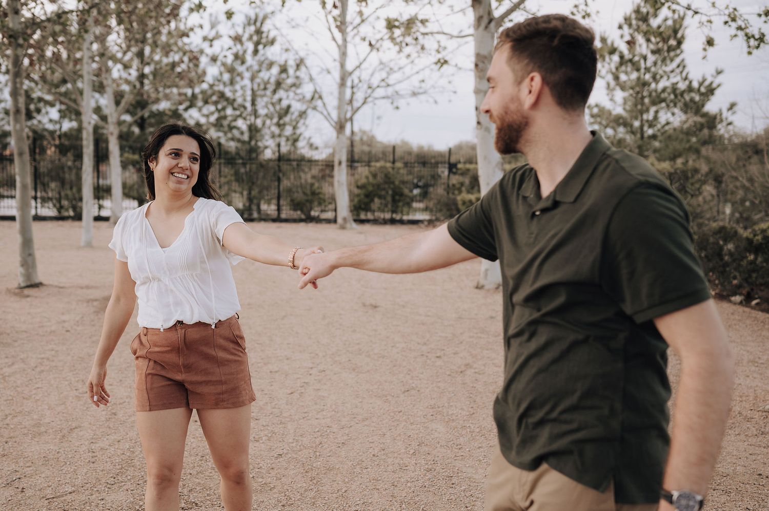 A playful couple reaching for each other's hands while dancing in an outdoor park.