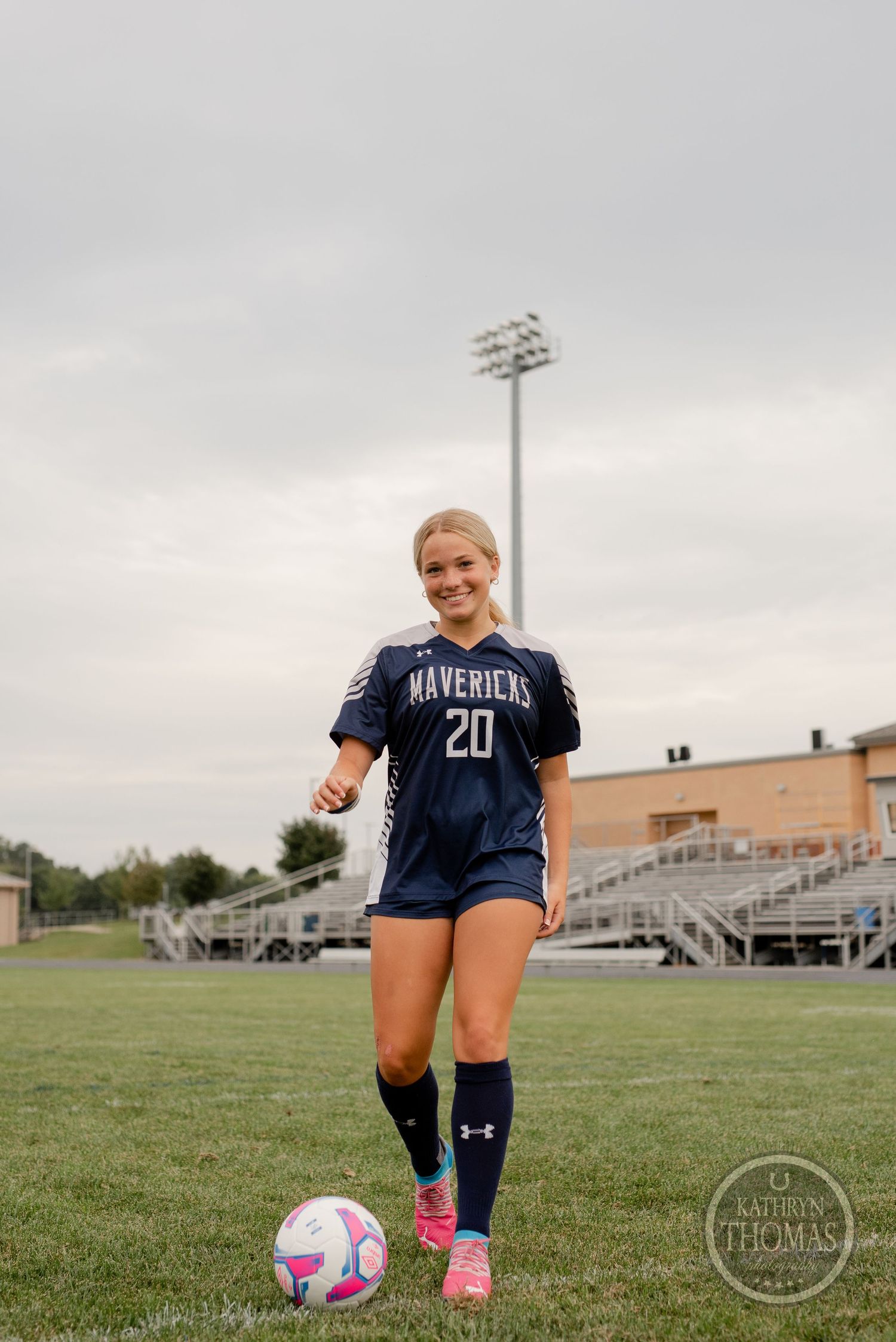 Soccer player in navy Mavericks uniform stands with ball on field under cloudy sky.