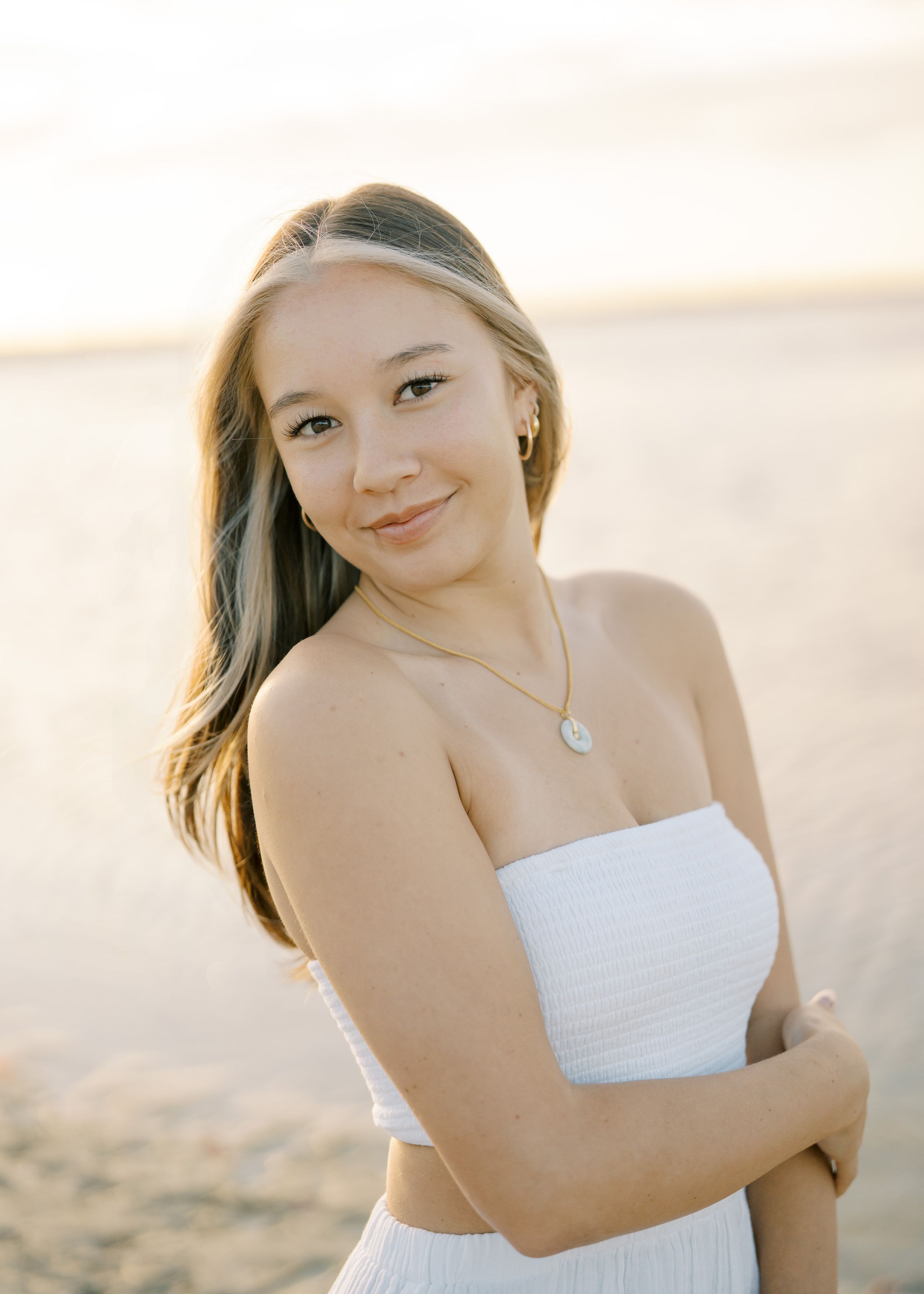 Teenage girl in white tube top smiling on sandy beach during golden hour sunset.