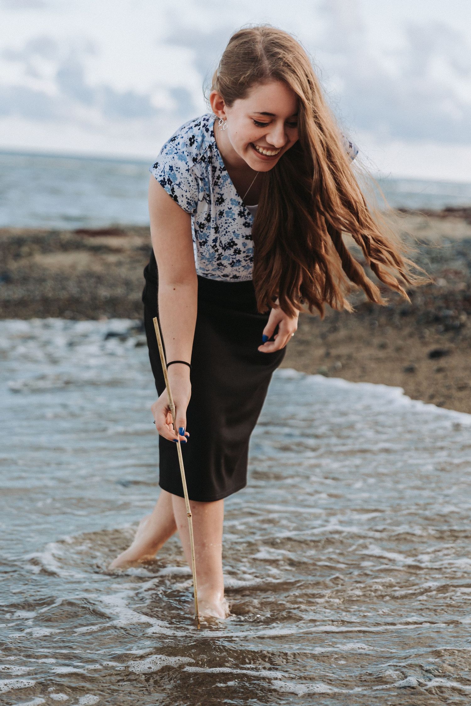 Woman in floral top and dark skirt playing in ocean waves with windblown hair.
