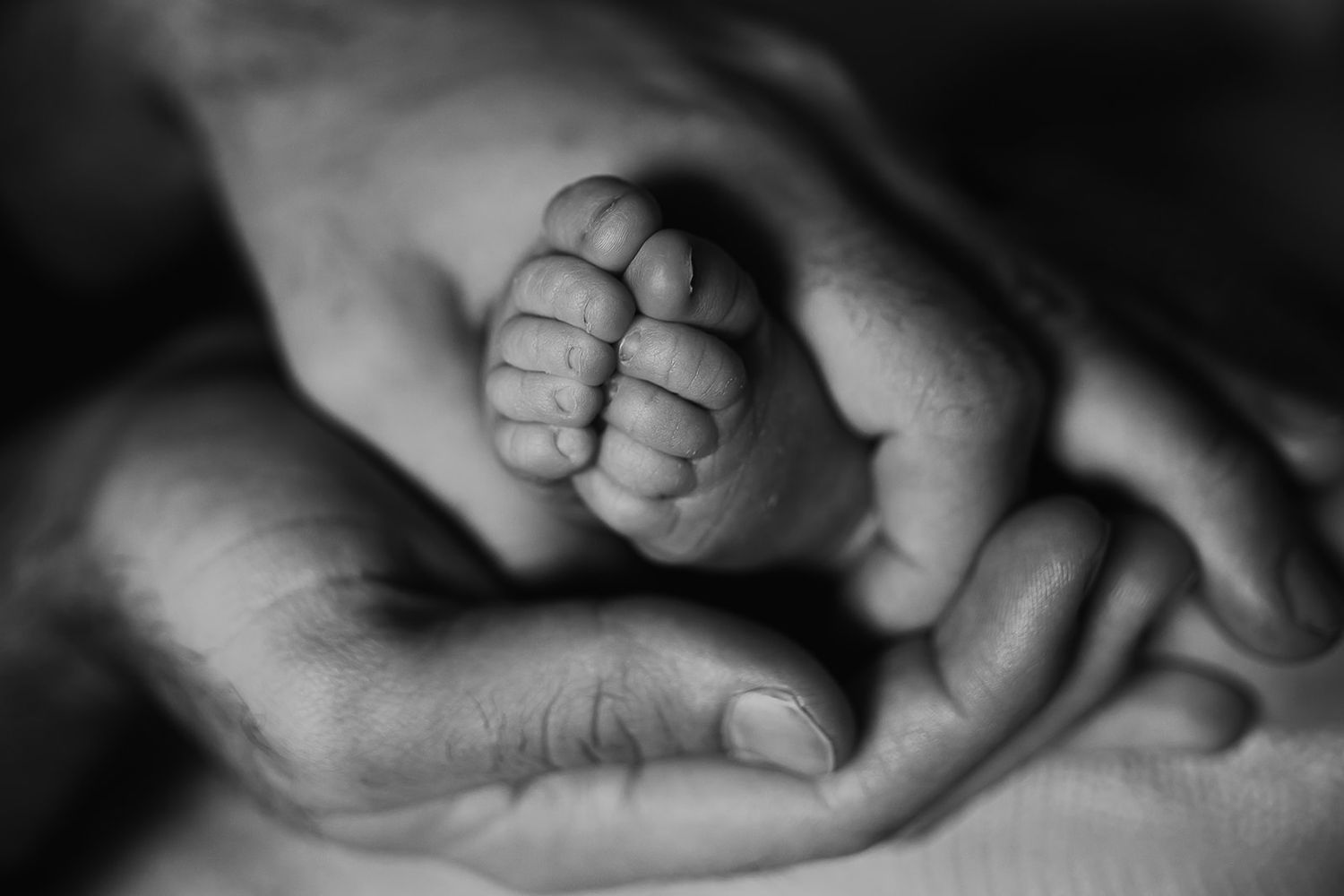 A black and white shot of a fathers hands wrapped around his newborn baby's feet.