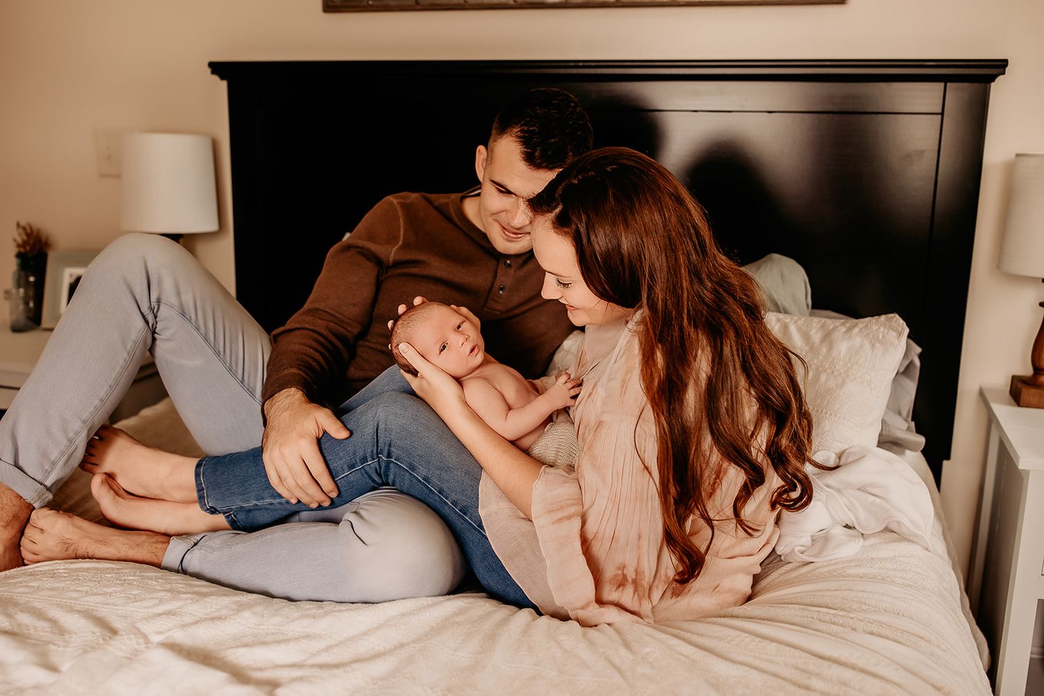 A couple sit on their bed smiling down at their baby boy.