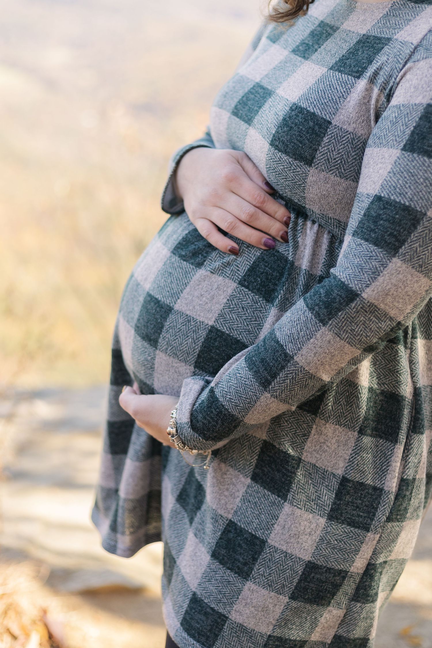 Close up of a plaid dress highlighting a baby bump against an autumn backdrop.