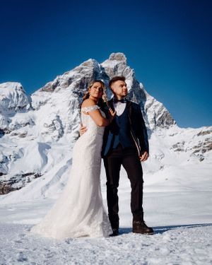 Couple poses in winter wedding attire against snow-capped mountain backdrop.