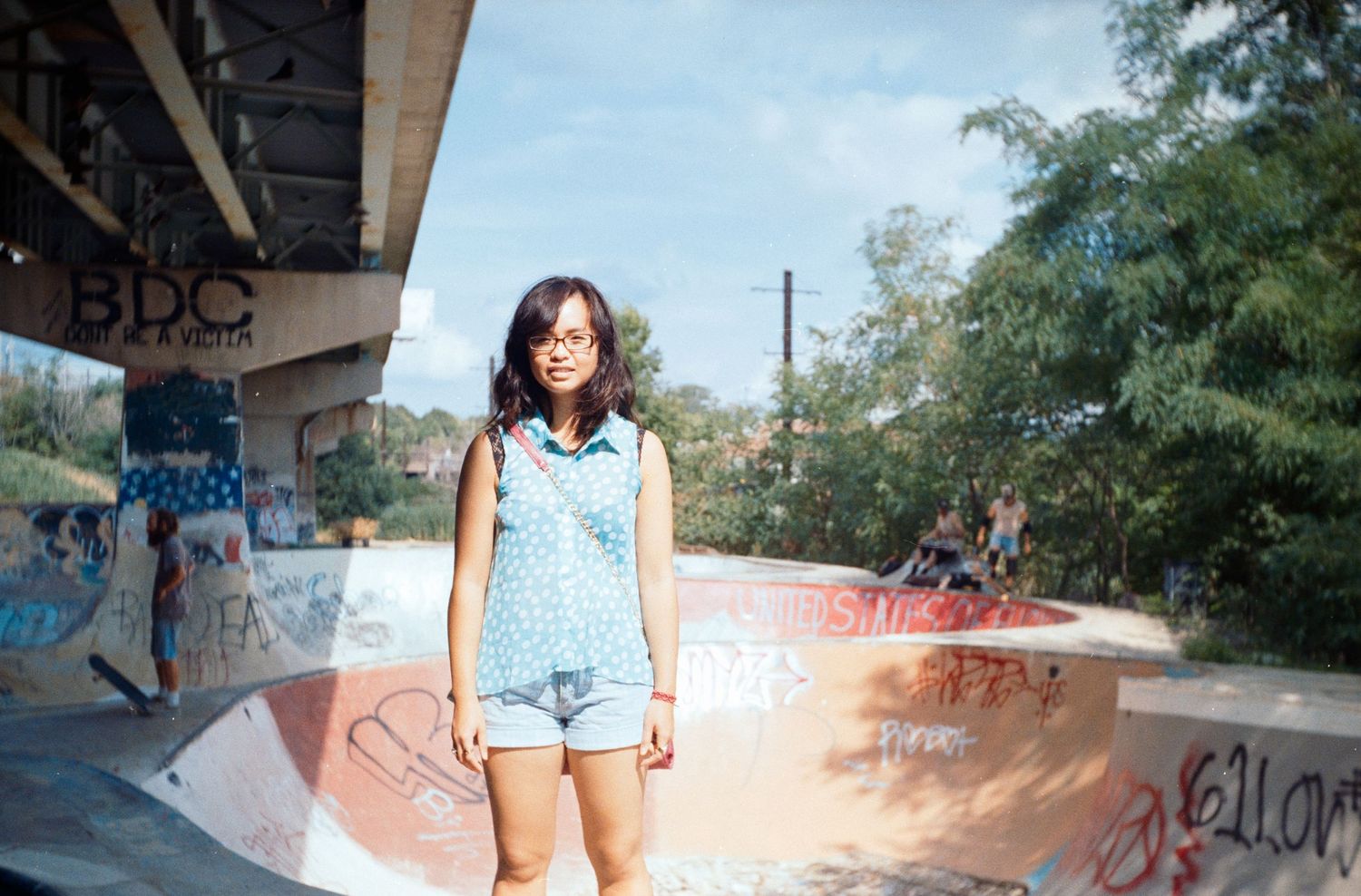 Graffiti-covered concrete skate park with ramps under bridge on sunny day.