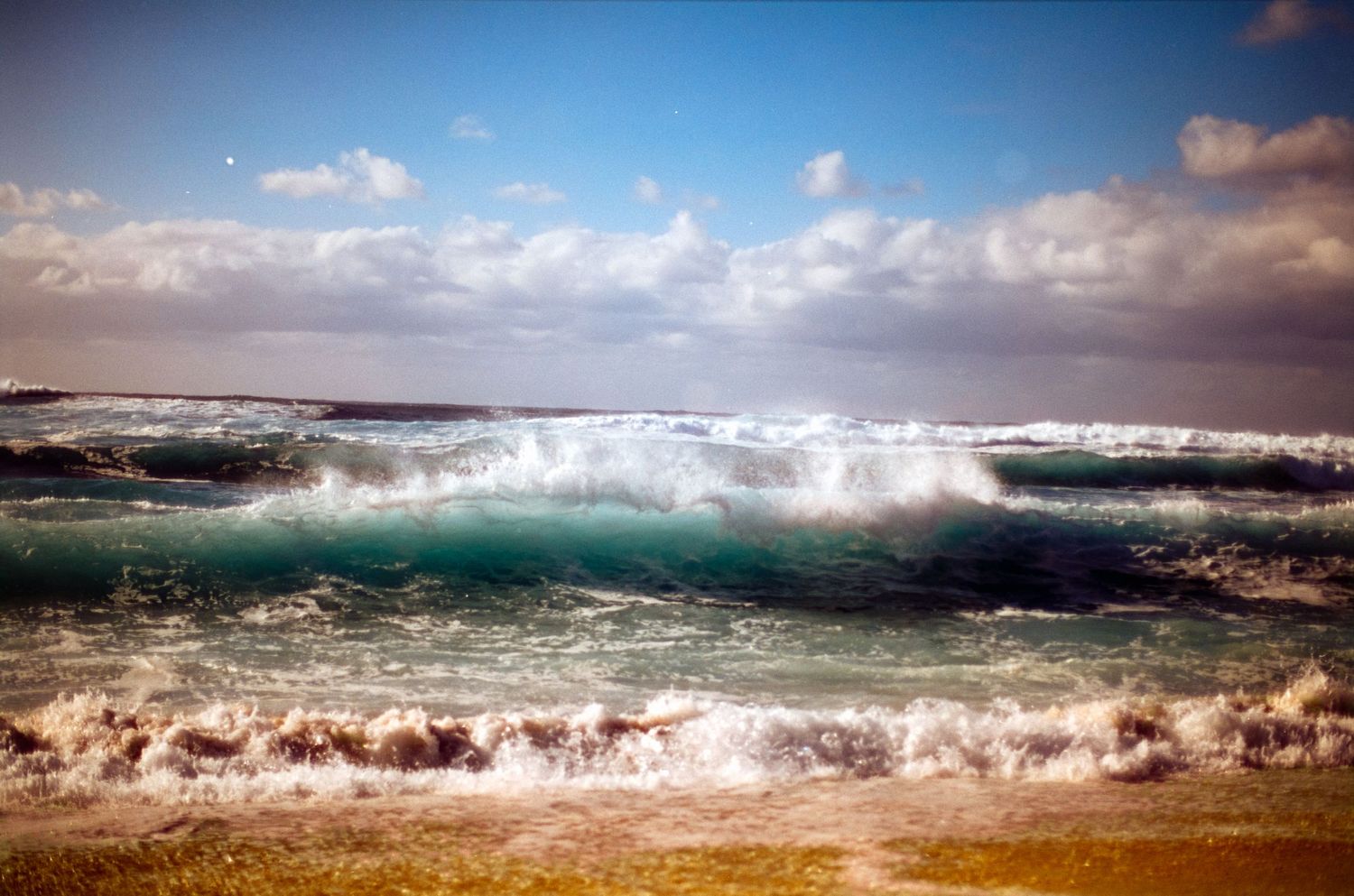 Dramatic ocean waves crashing on the shore with turquoise water under cloudy skies.