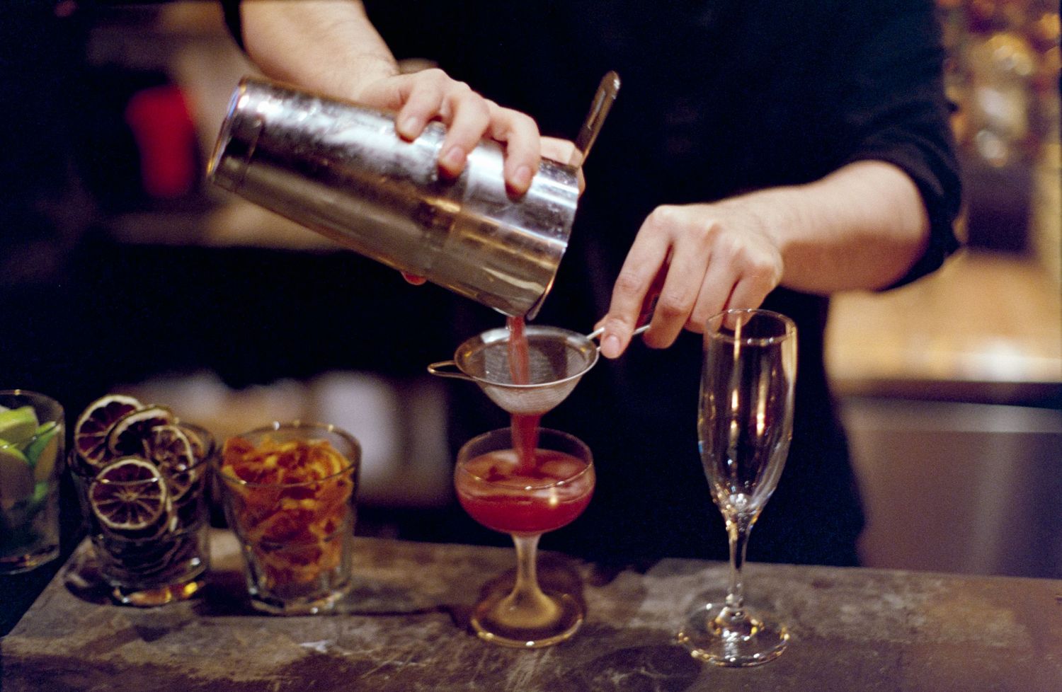 Hands pouring a cocktail from a shaker into a red martini glass.