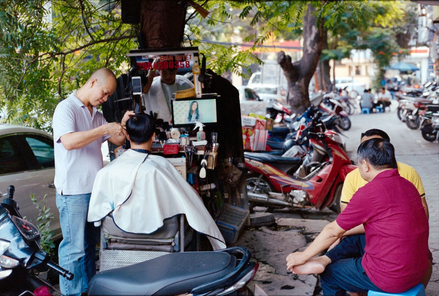 Street barber giving haircut outdoors with parked motorcycles in background on urban sidewalk.