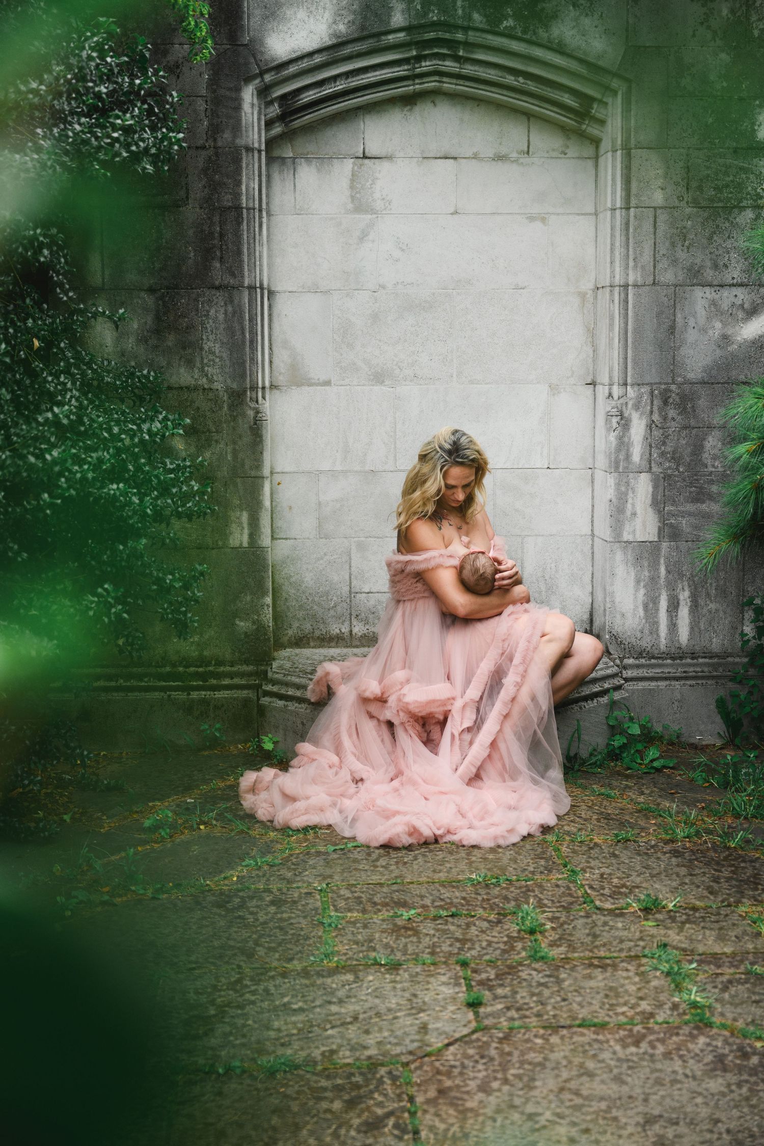 Woman in pink tulle gown cradling newborn baby outdoors near stone wall.