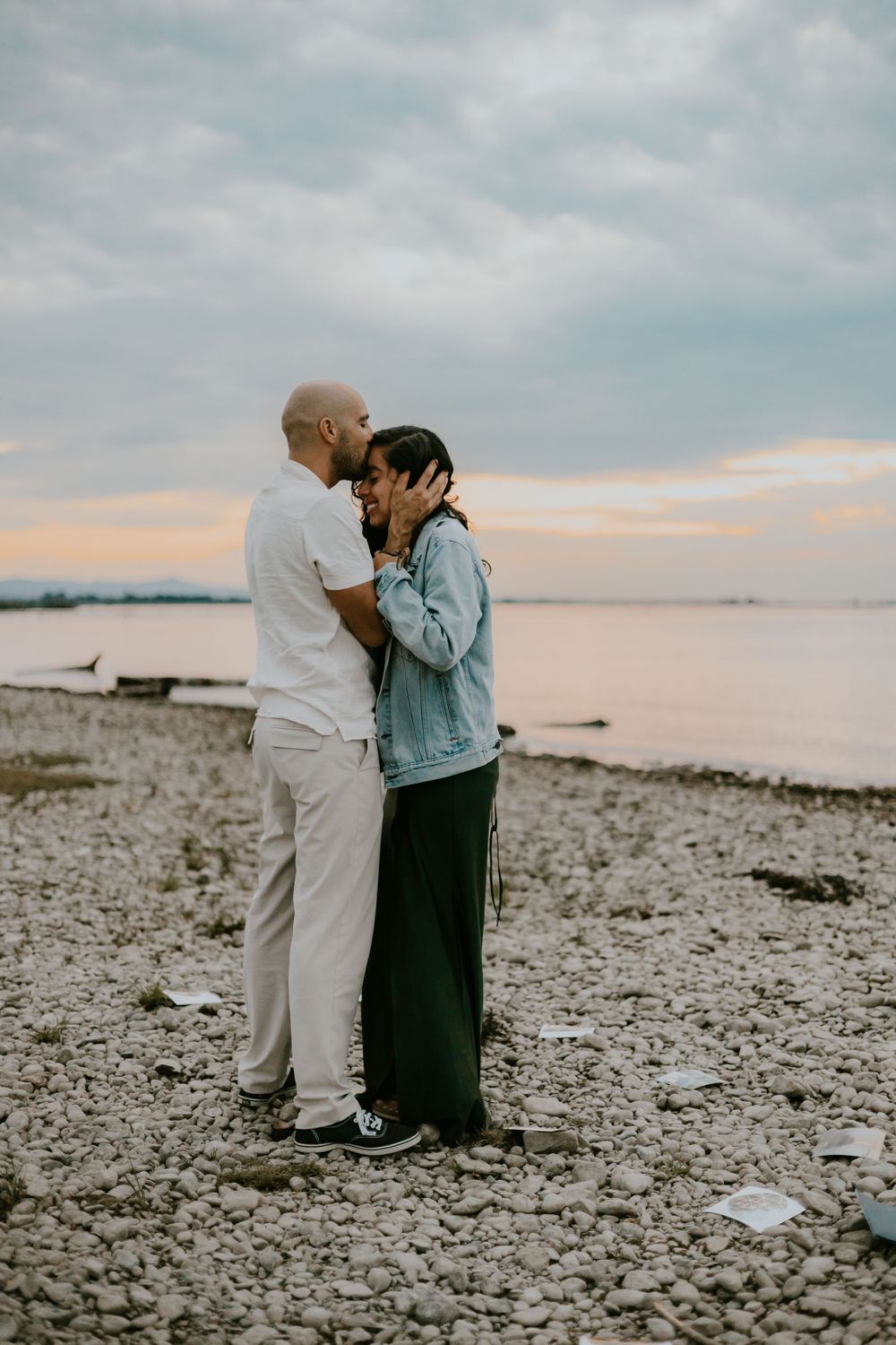 A romantic couple embraces on a rocky beach at sunset with one wearing a denim jacket and the other in white.
