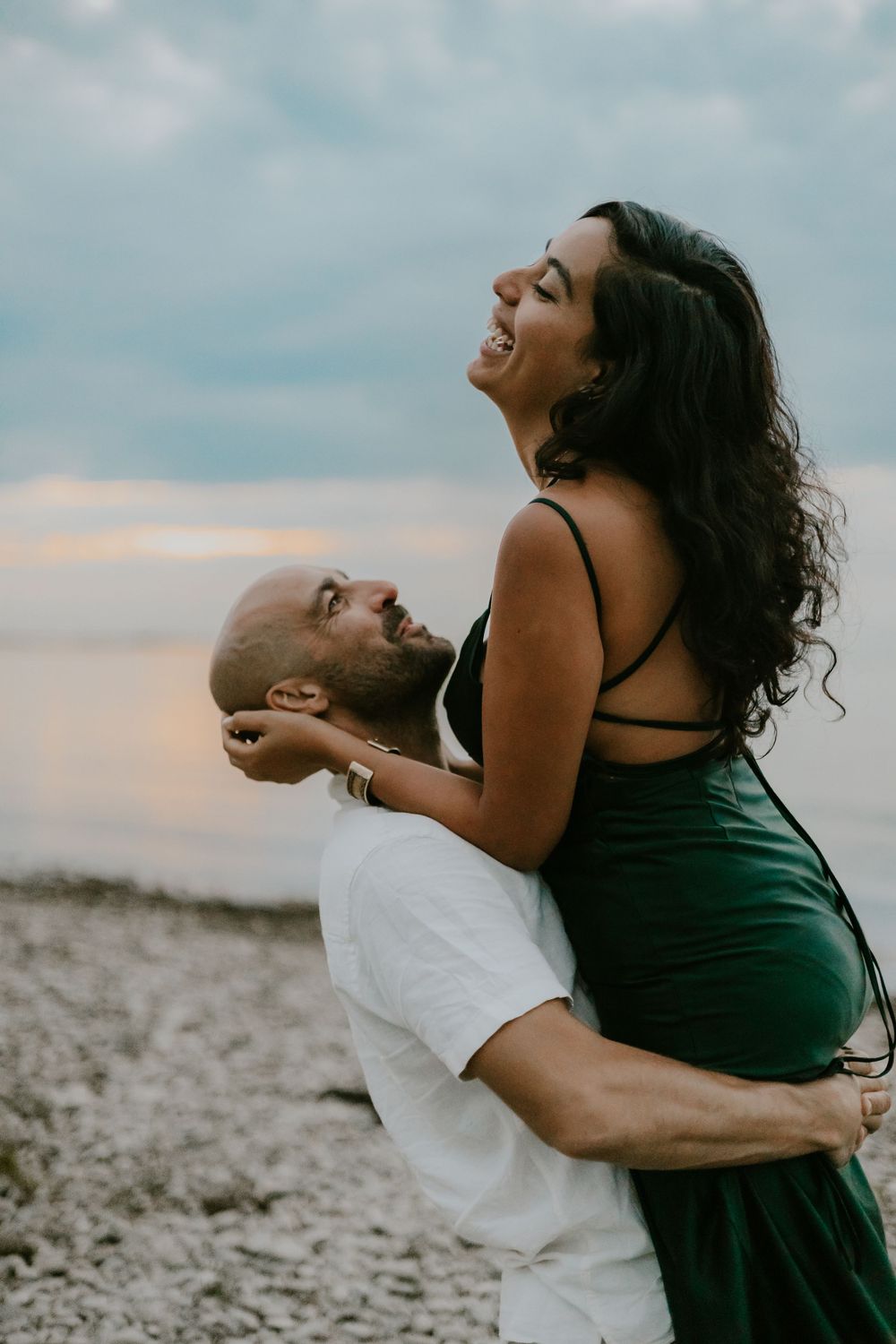 A playful couple shares a laugh while embracing on a rocky beach at sunset.