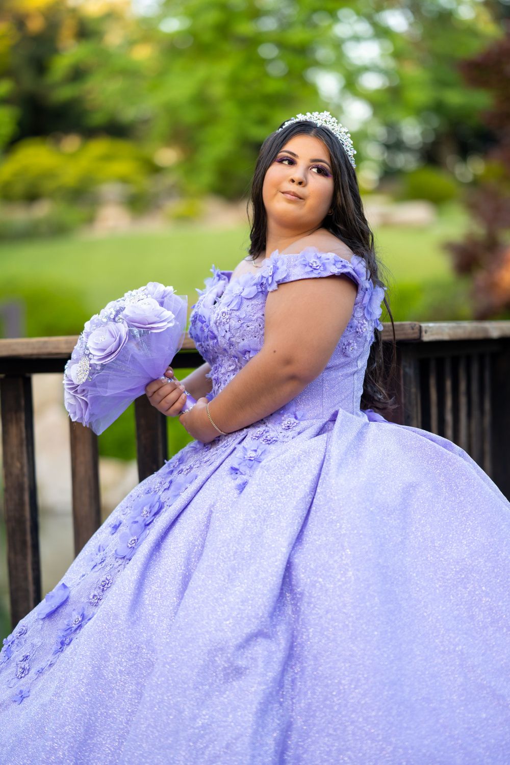 A person in a lavender quinceañera dress poses elegantly on a wooden deck in a garden setting.
