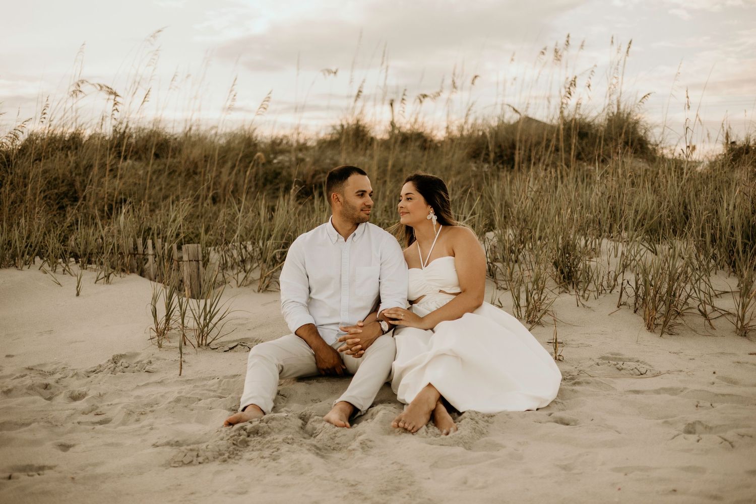 A couple sits together on sandy dunes at sunset in romantic beachwear.