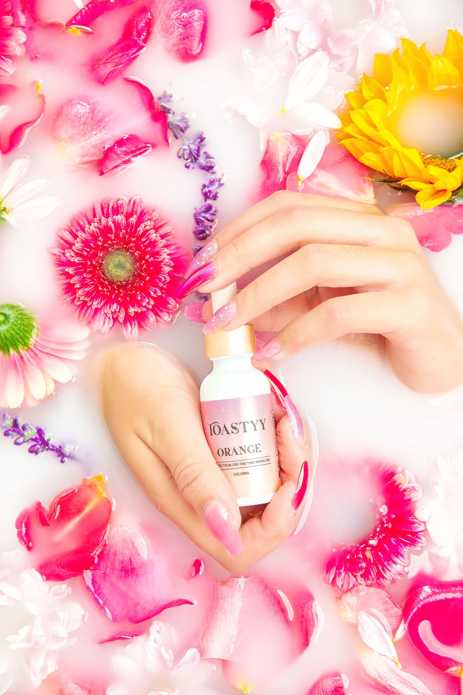 Hands holding small orange essential oil bottle surrounded by colorful flower petals in milky bath.