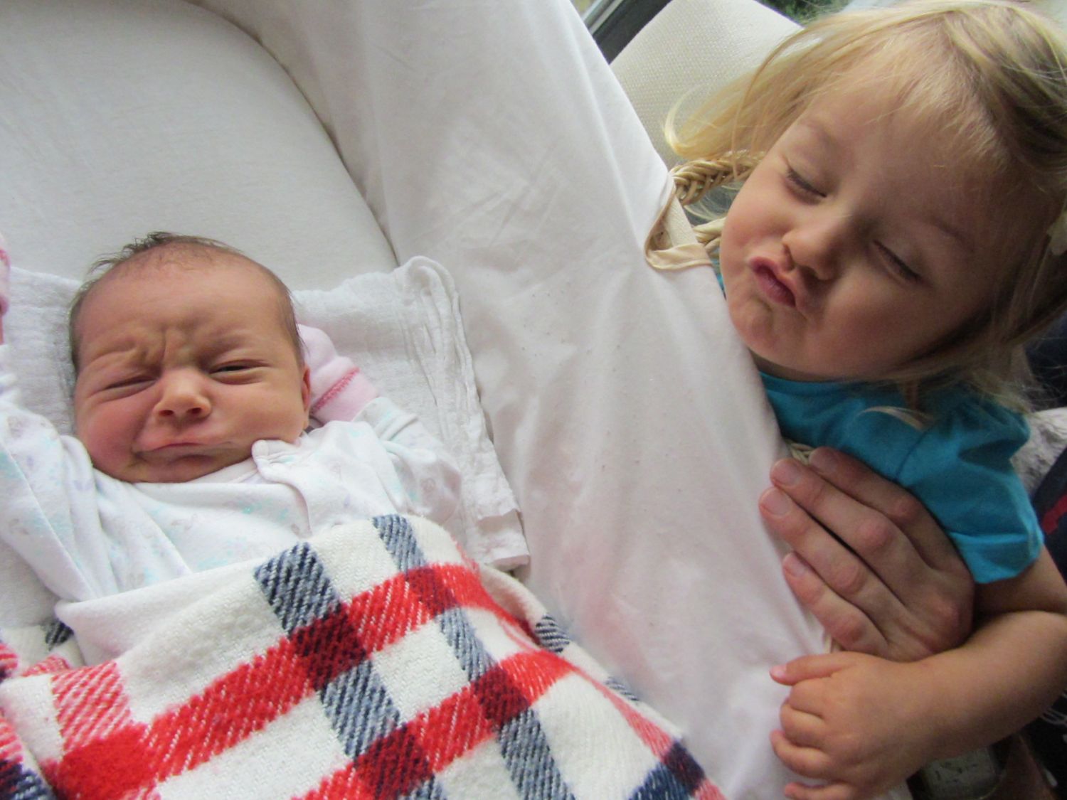 Two young children resting on white bedding, with a red and white plaid blanket.