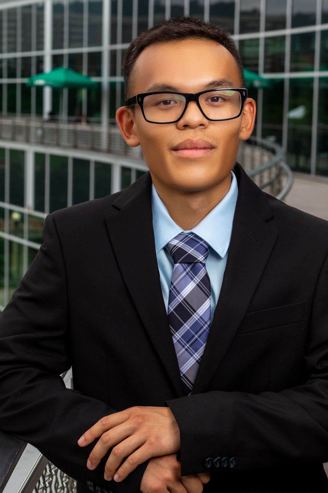 Professional headshot of young professional in black suit and glasses against modern office building exterior.