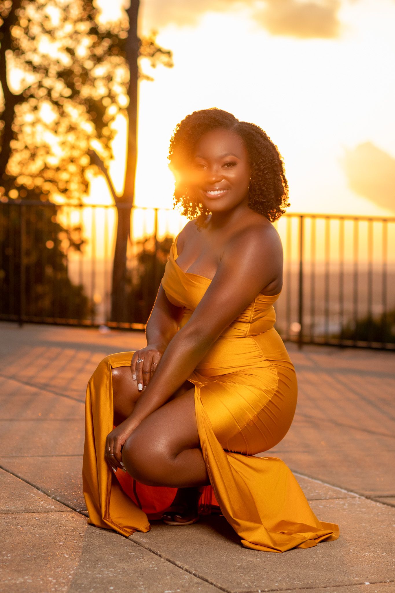 Woman in yellow evening gown poses during golden hour sunset on outdoor patio.