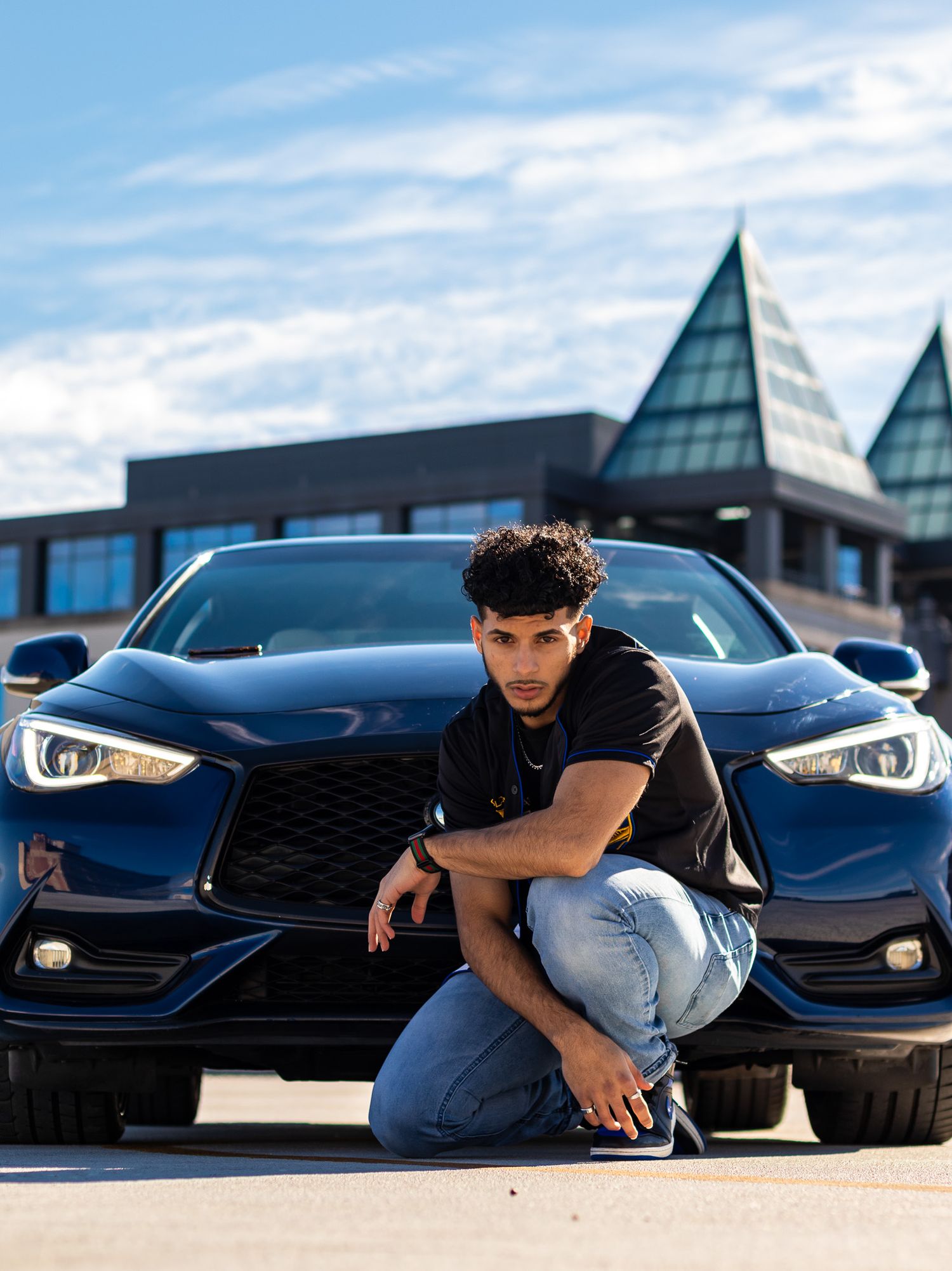 Casual portrait of person crouching next to black luxury car with modern architecture in background.