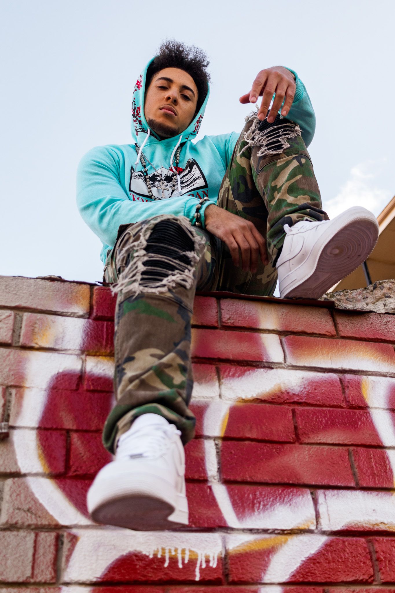 Urban fashion portrait of person in turquoise hoodie and camo pants sitting on graffitied brick wall.