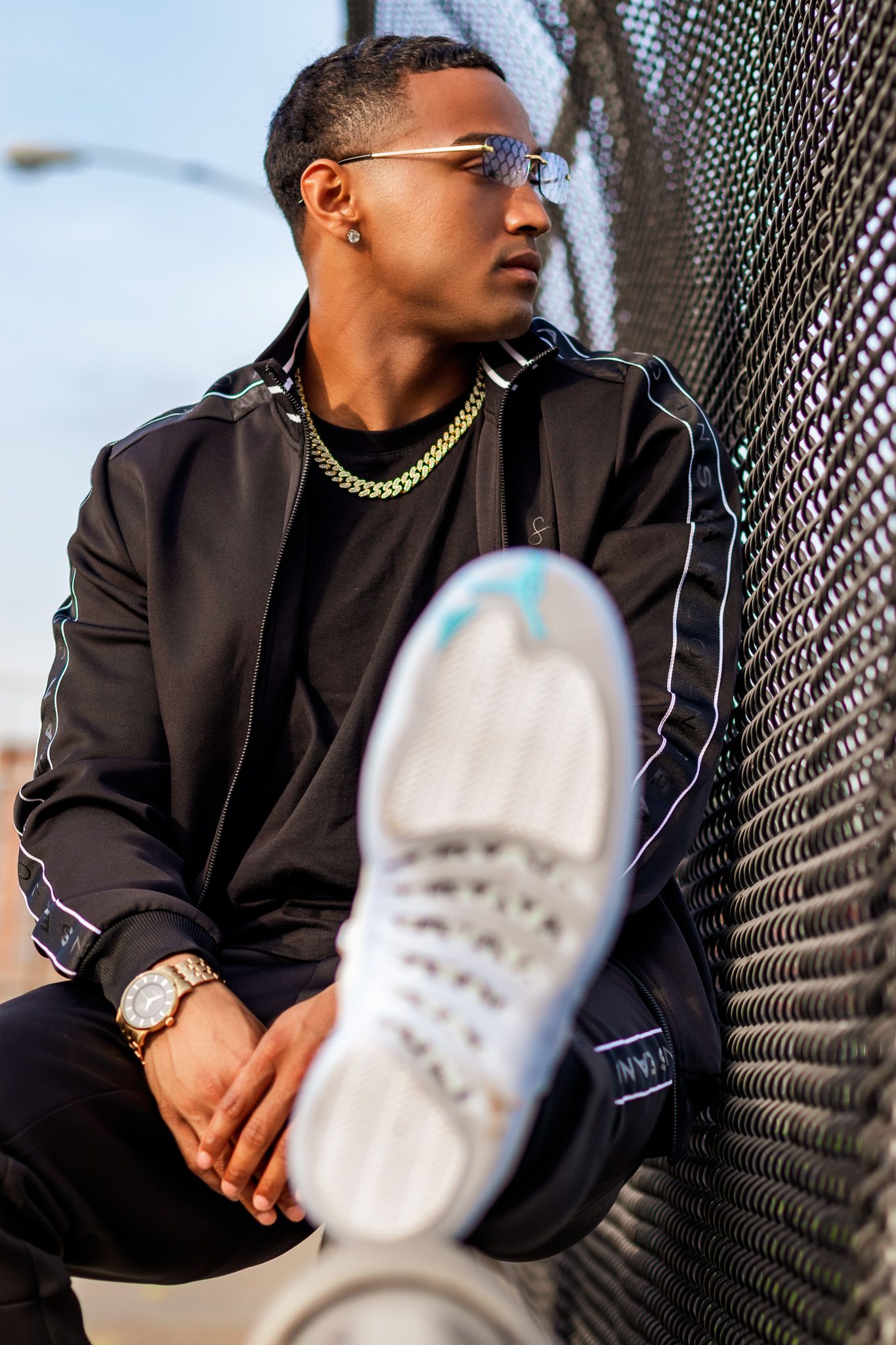 Stylish urban portrait of person in black jacket and gold chain against chain-link fence.