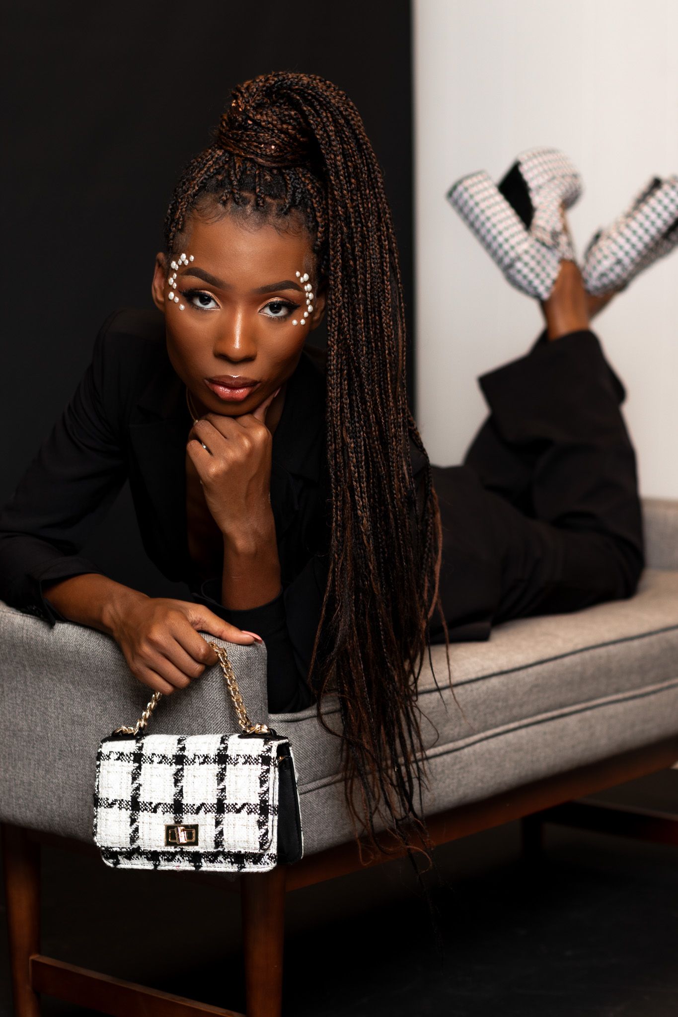 Fashion model with long braids poses on couch with black and white checkered handbag.