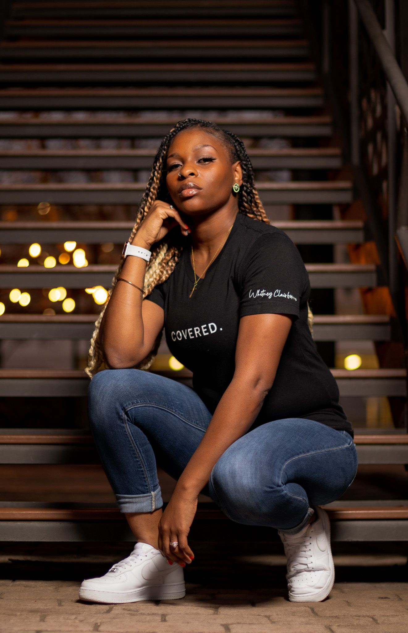 Stylish model in black t-shirt and jeans poses on wooden steps with warm lighting in background.