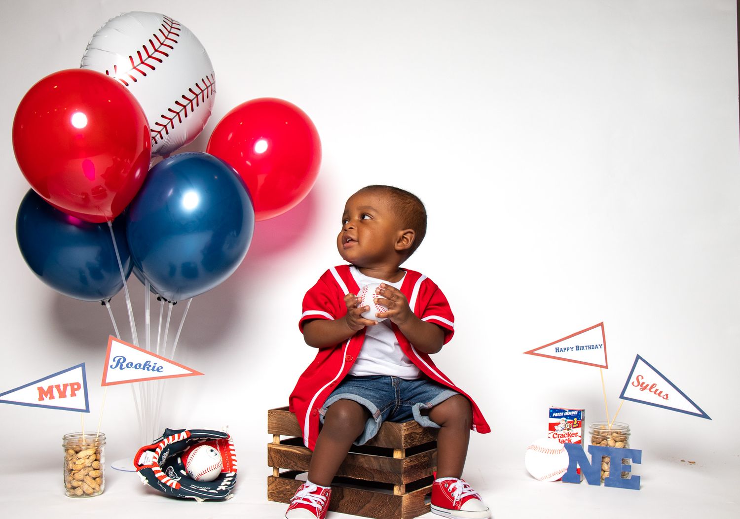 Baby in red outfit sits with baseball themed balloons and decorations for first birthday shoot.