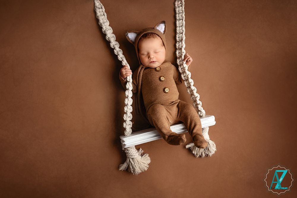 Sleeping infant in bear romper posed on decorative rope swing with white wooden seat.