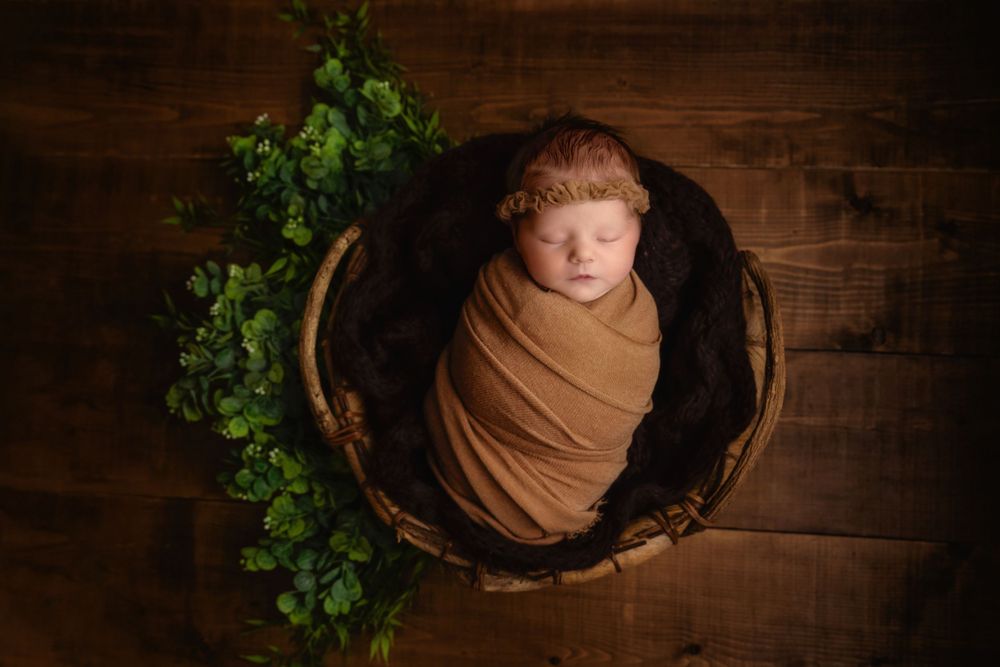 Sleeping newborn wrapped in brown swaddle lying in wooden bowl with green foliage decoration against rustic backdrop.