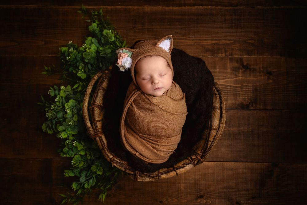 Peaceful newborn in brown swaddle with animal ears sleeping in wooden bowl with greenery accent.