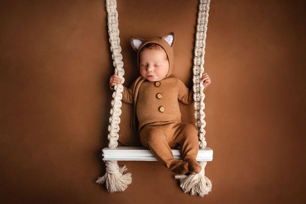 Baby wearing brown bear outfit resting on white macrame swing against mocha background.