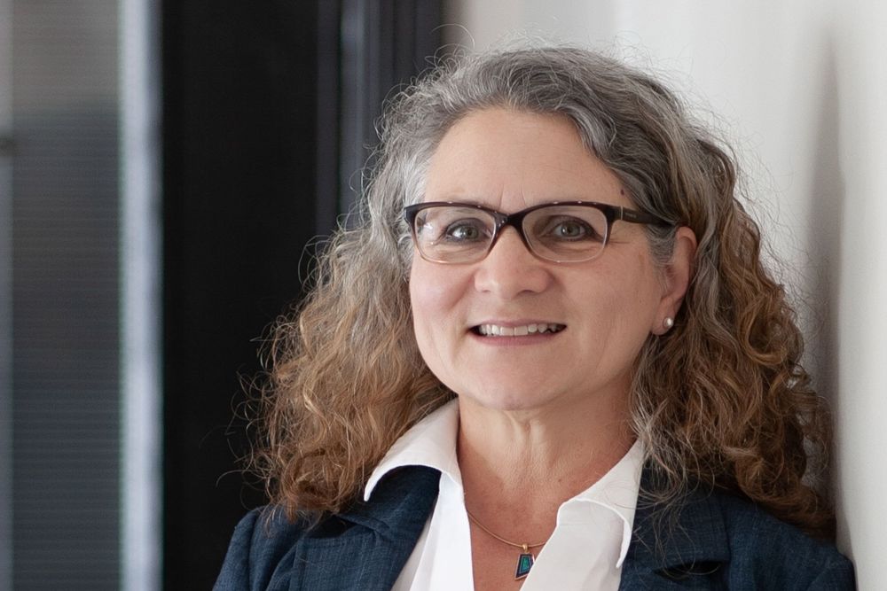 Professional woman with curly gray hair and glasses smiling warmly in a business casual setting.