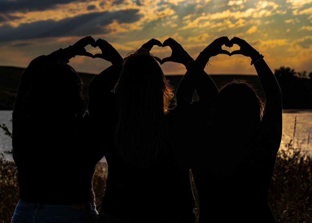 Silhouette of three friends making heart shapes with their hands against a dramatic sunset sky.