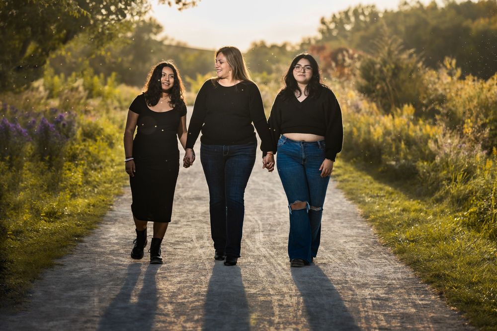 Three young women holding hands walking down a sunlit path surrounded by wildflowers at golden hour.