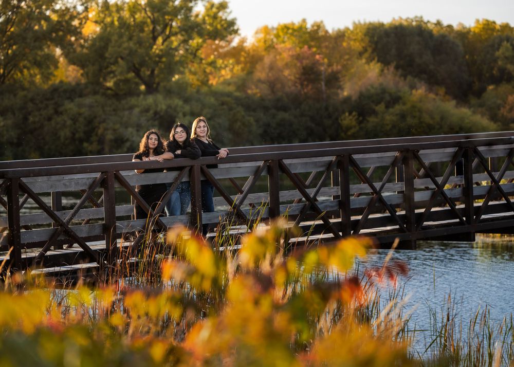 Three young women leaning on a wooden bridge railing surrounded by colorful autumn foliage at golden hour.