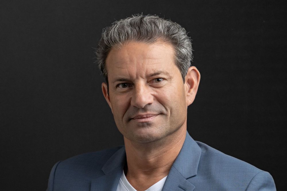 Professional headshot of man with gray hair wearing a blue blazer and white shirt against dark background.