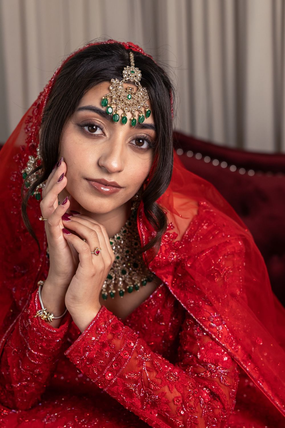Young South Asian bride in red bridal lehenga with gold maang tikka and emerald jewelry, posing elegantly.