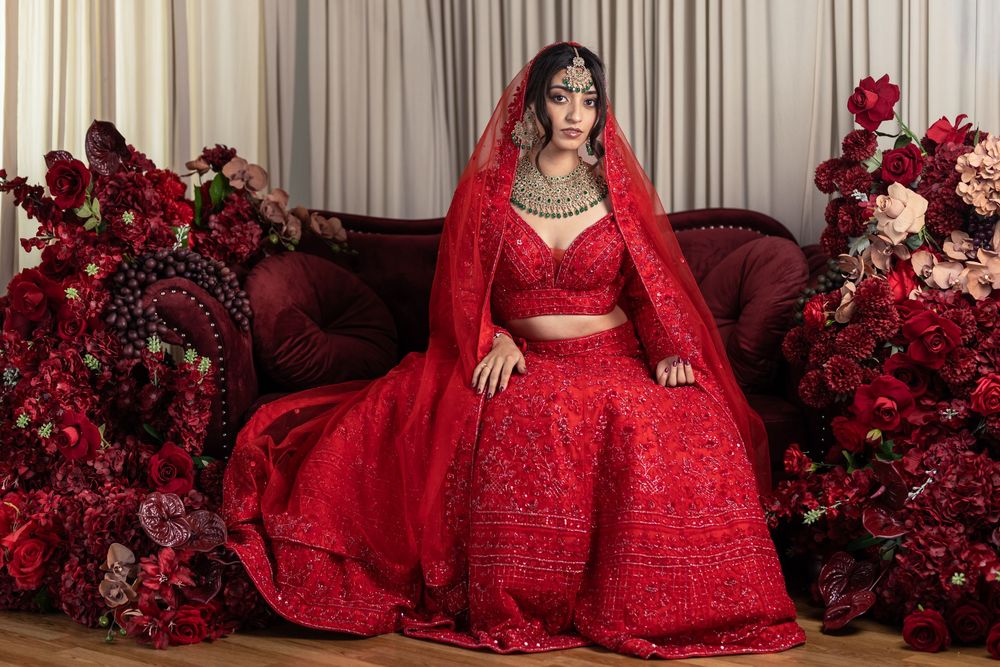 Bride in stunning red embellished lehenga with dupatta, seated among lush red floral arrangements in elegant bridal portrait.