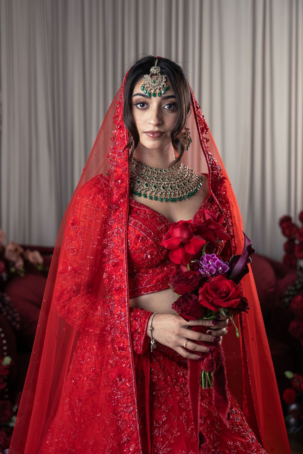 Indian bride in red lehenga with green gold jewelry holds red roses bouquet wearing traditional bridal dupatta.