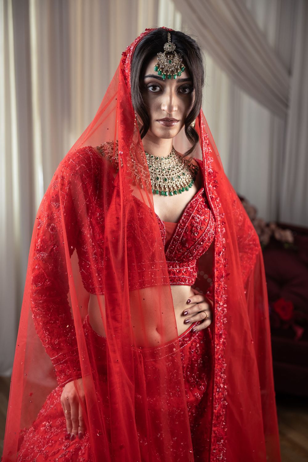 A bride in a red embroidered lehenga with veil, maang tikka, and gold kundan jewelry poses indoors.