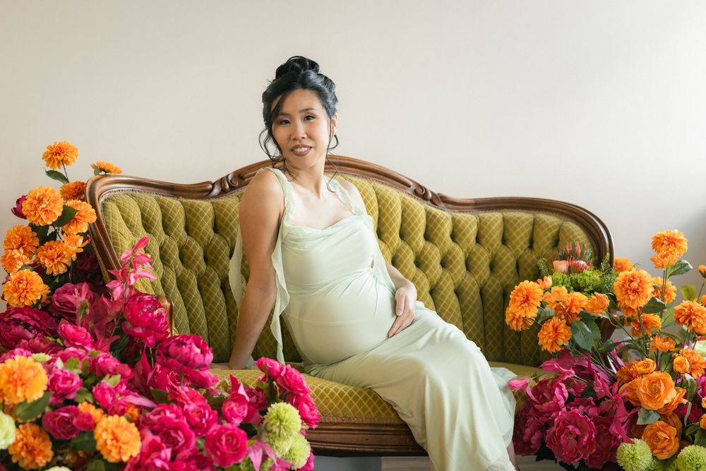 Pregnant woman in white dress sitting on green velvet sofa surrounded by colorful pink and orange flowers.