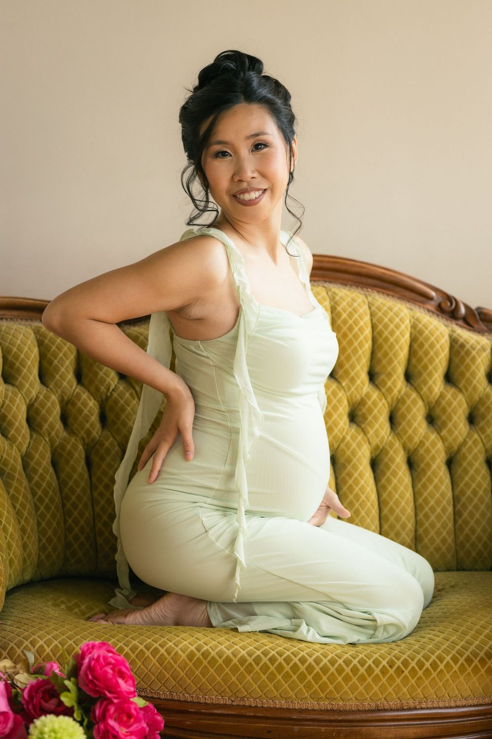 Smiling Asian woman in light green jumpsuit sitting gracefully on a vintage olive velvet sofa.