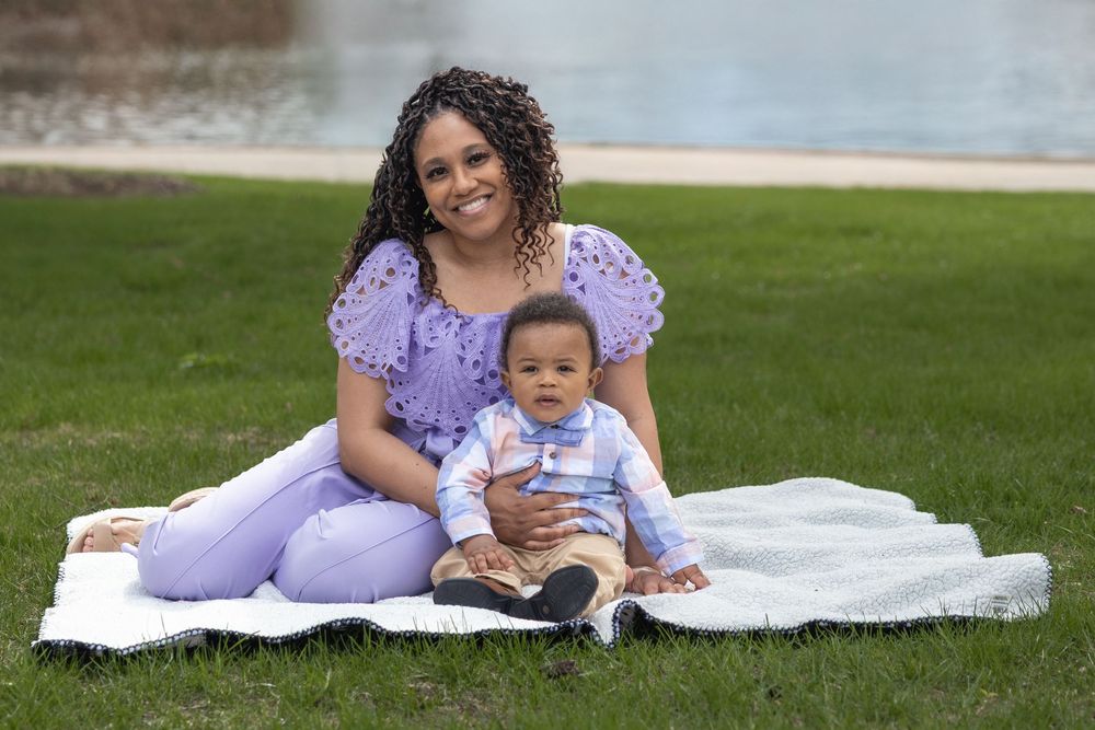 Smiling mother in purple outfit sitting on blanket in park with her baby boy.